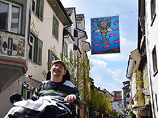Mann im Rollstuhl lächelt in einer sonnigen Altstadtstraße mit historischen Häusern, Bäumen und bunter Fahne, barrierefreies Stadtleben, Inklusion, behindertengerechte Mobilität, Freude am urbanen Alltag, Rollstuhlfahrer draußen