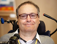 Mann mit Brille und grauem Pulli sitzt in einem elektrischen Rollstuhl, lächelt in die Kamera. Im Hintergrund unscharf ein buntes Bild. Perfektes Beispiel für barrierefreie Hilfsmittel für mobilitätseingeschränkte Menschen.