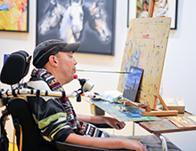 Mann im Rollstuhl malt mit einem Pinsel, den er mit dem Mund hält, ein farbenfrohes Bild auf einer Staffelei; barrierefreie Kunst, Inklusion und Kreativität, inspirierendes Künstlerbild im Kunststudio mit Gemälden im Hintergrund.