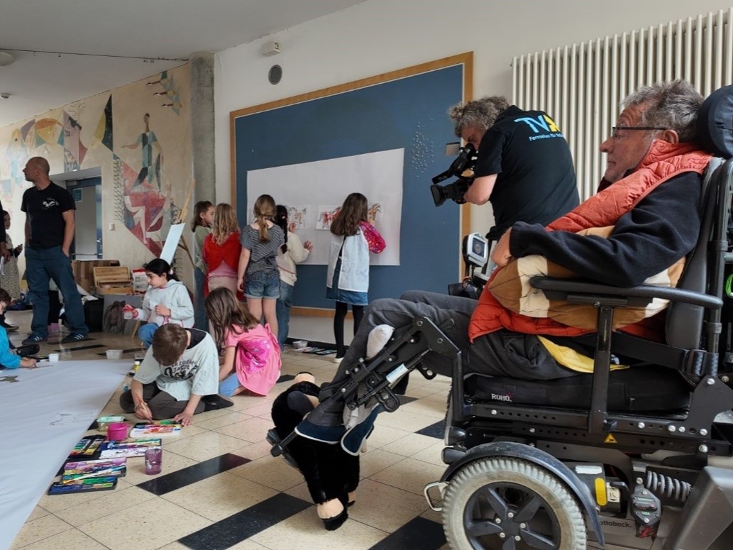 Mundmaler Markus Kostka demonstriert in einem Klassenzimmer der Schule ViTo sein handwerkliches Können: Mit dem Mund hält er einen Pinsel und malt auf einem großen weißen Papier, das auf einer blauen Tafel befestigt ist. Eine Gruppe von etwa zehn Grundsch