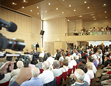 Großer, heller Konferenzraum mit vielen sitzenden und stehenden Menschen, ein Redner spricht am Podium, Zuschauer auf Empore, Kamera im Vordergrund filmt, Veranstaltung oder Vortrag, professionelle Event-Atmosphäre.