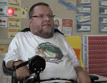 Mann im Rollstuhl mit Brille und weißem T-Shirt mit Tiermotiv sitzt in einem bunt dekorierten Klassenraum, umgeben von Lernpostern und Tafeln für Kinder, Fokus auf Inklusion, Barrierefreiheit und Bildung.