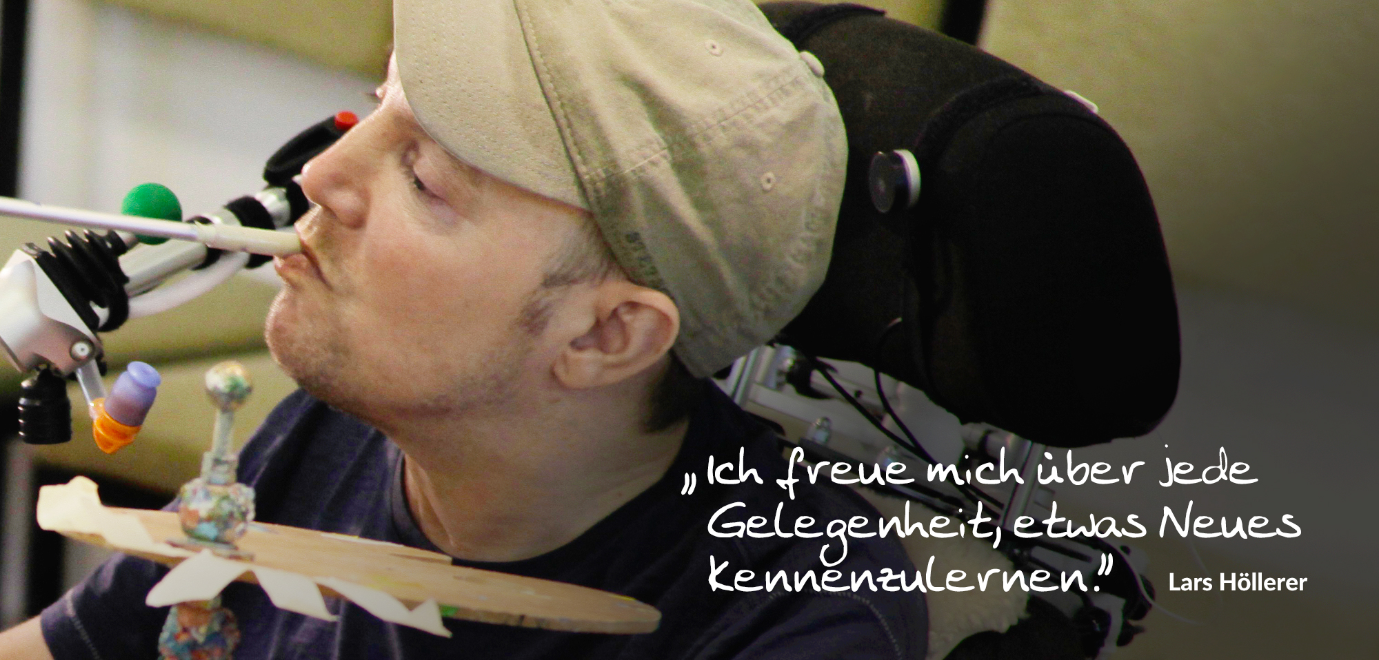 Mann im Rollstuhl mit Basecap nutzt eine Mundsteuerung zum Malen, Farbpalette und Pinsel sichtbar; Zitat: „Ich freue mich über jede Gelegenheit, etwas Neues kennenzulernen.“ Perfektes Hilfsmittel für barrierefreie Kreativität.