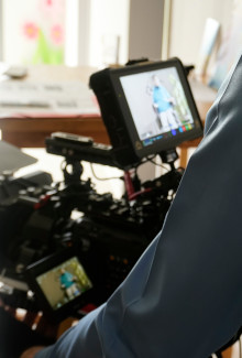 Professionelle Filmkamera mit großem Monitor und Bildschirm zeigt bei Tageslicht im Innenraum eine Person, unscharfer Hintergrund mit Tisch und Unterlagen, hochwertige Videoausrüstung für Filmproduktion oder Videodreh.