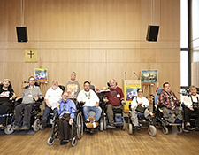 Gruppe von zehn Menschen mit unterschiedlichen Behinderungen in Rollstühlen in einem barrierefreien, hellen Raum mit Holzverkleidung; inklusives Miteinander, Diversität, Gemeinschaft, Barrierefreiheit, Teilhabe, soziales Engagement.