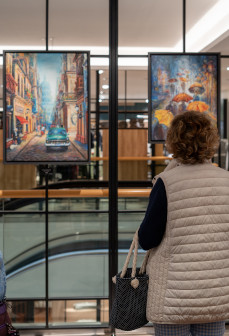 Frau mit hellbeiger Steppweste und Handtasche betrachtet in modernem Einkaufszentrum zwei bunte Wandbilder; links Stadtstraße mit Oldtimer, rechts Regenschirme – ideal als dekorative Wandbilder für stilvolle Wohnräume.
