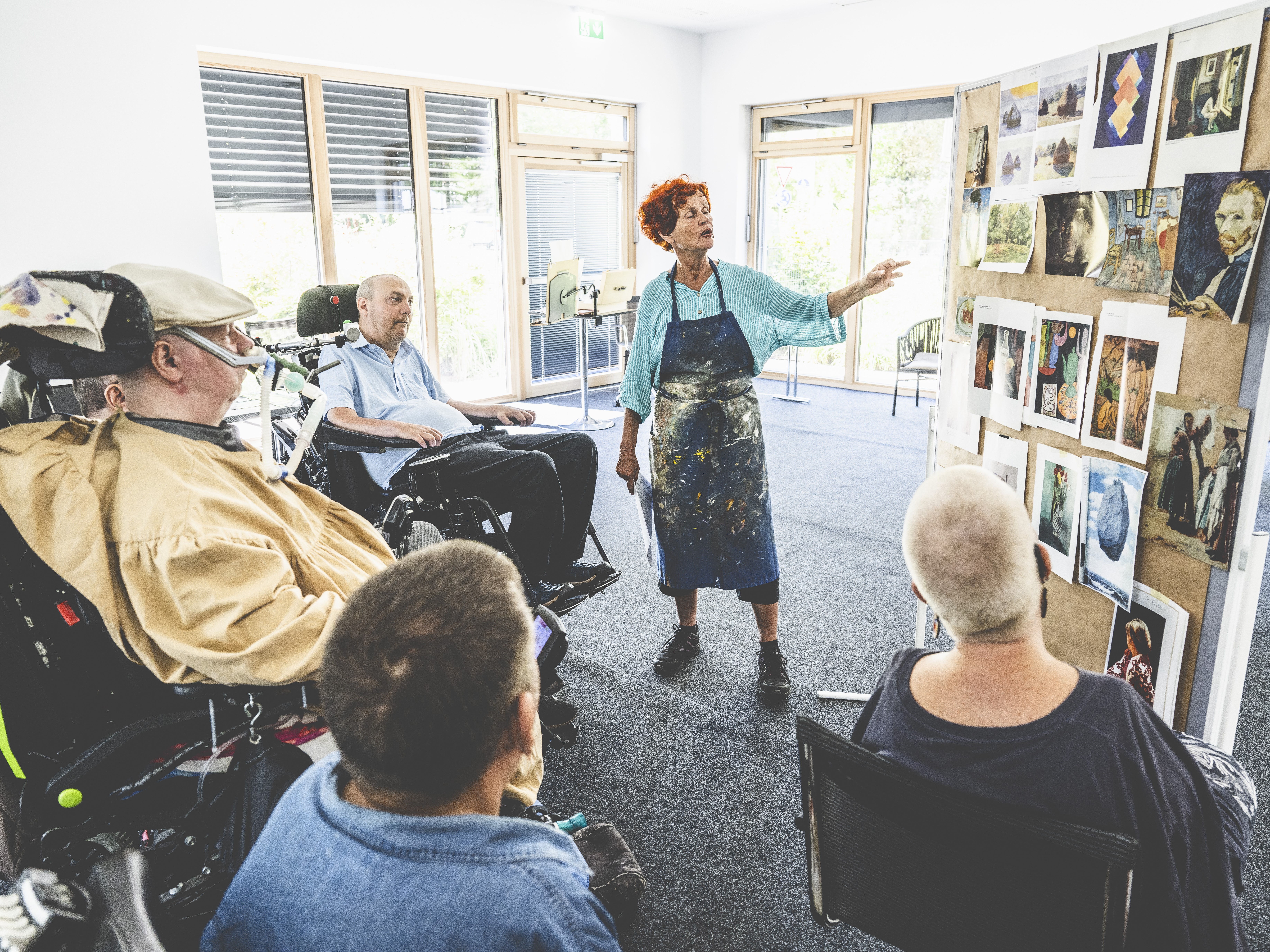 Künstlerin erklärt Menschen mit Behinderung im Rollstuhl verschiedene Kunstwerke auf einer Pinnwand in hellem Kreativraum, inklusive Kunstworkshop fördert Barrierefreiheit, Kreativität und Teilhabe im Alltag für alle Teilnehmer.
