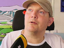 Mann im Rollstuhl mit beigem Cap und weißem Shirt sitzt vor farbigem Hintergrund, hält gelbes Mikrofon, barrierefreie Kommunikation im Alltag, Inklusion, Assistenzbedarf, Produkt für Menschen mit Behinderung, Onlineshop.