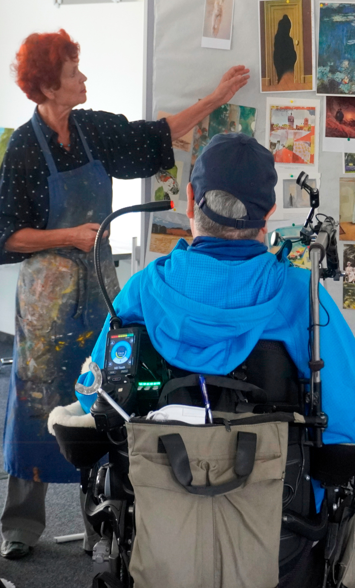 Künstlerin erklärt einer Person im elektrischen Rollstuhl mit Kommunikationsgerät verschiedene Kunstwerke an einer Pinnwand, inklusive Malereien und Fotografien, in einem inklusiven Kunstworkshop für Barrierefreiheit und Teilhabe.