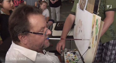 Mann mit Bart malt ein Bild mit dem Mund an einer Staffelei, Zuschauer beobachten aufmerksam. Barrierefreies Malen, Inklusion, Kunst für Menschen mit Behinderung, Kreativität ohne Hände, Maltechniken für Menschen mit Einschränkungen.