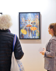Zwei Personen betrachten in einer hellen Galerie ein farbenfrohes Gemälde mit Menschen und bunten Regenschirmen. Das Bild ist gerahmt und hebt sich lebendig vor der weißen Wand hervor – ideal für Kunstliebhaber und moderne Wohnräume.