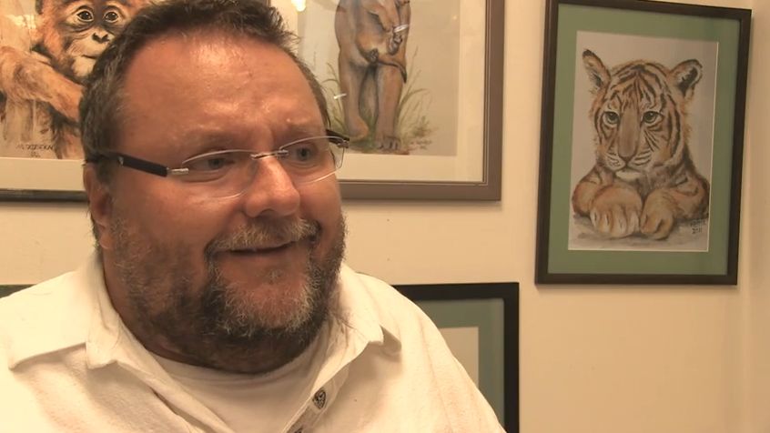 Mann mit Brille und Bart lächelt freundlich in die Kamera, trägt weißes Hemd; im Hintergrund Gemälde von Tieren wie Tiger und Affe an weißer Wand, ideal für Kategorie Tierporträts Kunstdrucke Wanddeko.