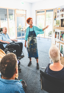 Künstlerin präsentiert kreativen Workshop vor mehreren Personen, darunter ein Mann im Rollstuhl. Die Gruppe sitzt aufmerksam vor einer Pinnwand mit Bildern in einem hellen, barrierefreien Raum mit großen Fenstern.