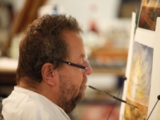Mann mit Brille malt am Arbeitsplatz mit dem Mund einen farbenfrohen Bildausschnitt auf eine Leinwand, konzentrierter Ausdruck, barrierefreie Kunst im Atelier, inklusive Kreativität, Künstlerbedarf online kaufen.