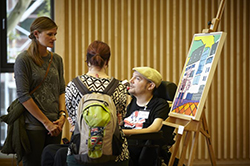 Drei Menschen betrachten gemeinsam ein farbenfrohes Gemälde auf einer Staffelei, darunter ein Mann im Rollstuhl und zwei Frauen, Gespräch und inklusive Kunst im modernen, barrierefreien Raum.