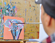 Künstler malt mit dem Mund ein buntes Elefantenbild auf Leinwand vor Staffelei, unscharfer Hintergrund und Maler im Vordergrund, kreative Mundmalerei, barrierefreie Kunst für Onlineshop, integrative Kunstprodukte